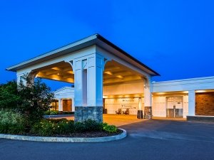 Evening view of a hotel's covered entrance with a circular driveway and landscaped greenery. The building features white columns, stone accents, and a modern design.