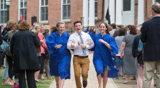 Three Saint Lawrence students run down a side walk and laugh as a crowd watches on. Two students are wearing vibrant blue robes.