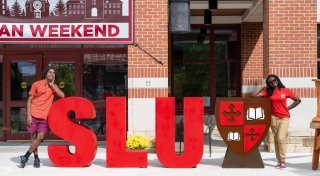 Two Saint Lawrence University students stand on each side of large S, L, U letters. They are wearing bright red polos and standing outside of the Sullivan Student Center on a sunny day.