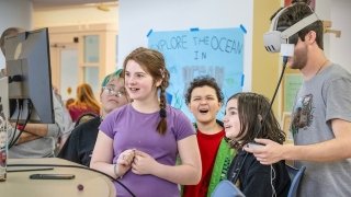 A group of kids excitedly watch as a person uses a virtual reality headset in a classroom setting. A sign in the background reads “Explore the Ocean.” One child holds a controller while others smile and laugh.