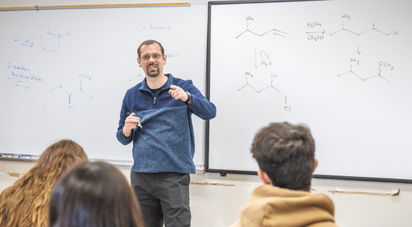 Sam Tartakoff teaches chemistry, gesturing while explaining concepts on a whiteboard filled with chemical structures. Students sit in the foreground, listening attentively.