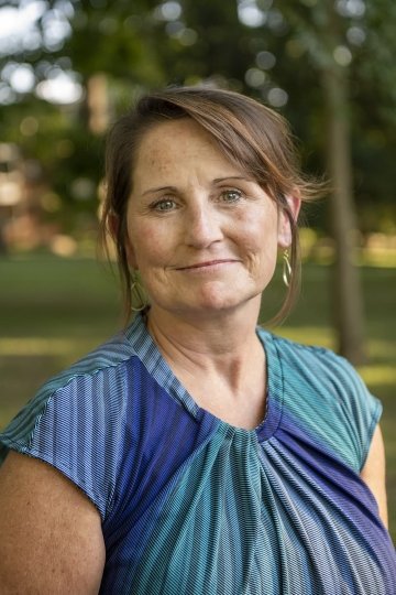 Headshot of Karen Butler, standing against a backdrop of trees. Her hair is partially pulled back and she wears a patterned shirt with many shades of blue and green in it.
