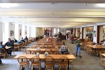Dana Dining Hall filled with students eating, studying, or conversing. Natural light streams in from tall windows on the left, illuminating the open seating area. In the back, a food service area with additional seating is visible.