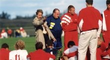 Bob Durocher, in a tan trench coat, is giving instructions to his team during a break in a game. The coach is gesturing with his hand while the players, dressed in red and white uniforms, sit and listen attentively. 
