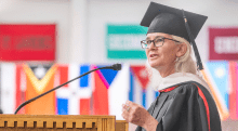 A person in a graduation cap and gown stands at a wooden podium, speaking into a microphone. The individual, who appears to be a faculty member or guest speaker, has white hair and wears glasses. In the background, colorful flags representing various countries or institutions are slightly blurred, creating a vibrant backdrop for the scene. The speaker's expression is focused and engaging, emphasizing the importance of the occasion.