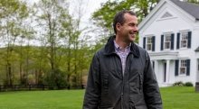 Brian Spitzmiller, wearing a dark green barn coat, stands smiling in a lush green yard. A large white house is in the background.
